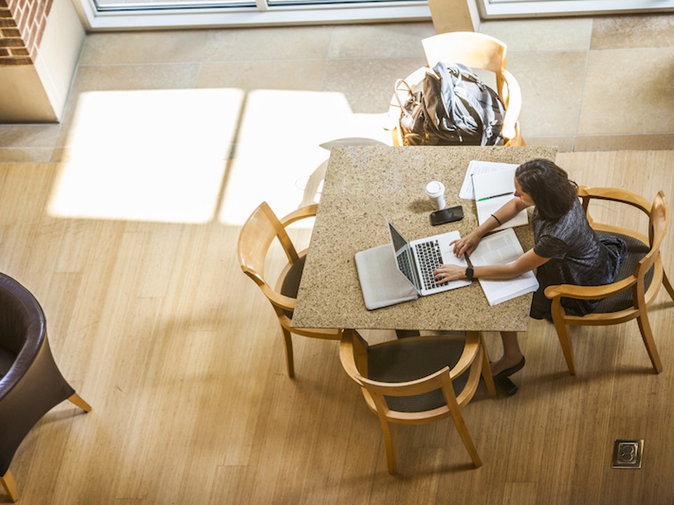 A female Online MBA student sits at a brown table working on her laptop while surrounded by papers and books